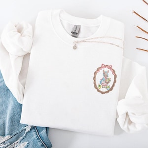 Könnte beinhalten: Weißes Sweatshirt mit Rundhalsausschnitt und gesticktem Oster-Design. Das Design zeigt ein Häschen in einem Kleid, das einen Korb mit Eiern hält, eingerahmt in einem Oval mit einer rosa Schleife. Eine silberne Halskette mit einem kleinen Anhänger ist ebenfalls sichtbar.