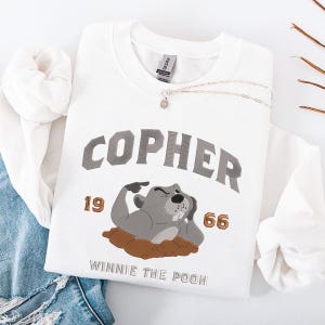 May include: White sweatshirt featuring the word "COPHER" in grey arched lettering, a cartoon gopher illustration, and the text "WINNIE THE POOH". The sweatshirt also features the numbers "19" and "66" in a gold colour. A silver necklace with a pendant is also visible.