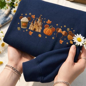Peut inclure: Un sweat-shirt bleu marine orné de broderies sur le thème de l'automne, incluant une boisson à la citrouille, un château, une citrouille, des oreilles de souris avec un nœud et un gland, entourés de petites étoiles et de feuilles d'érable.