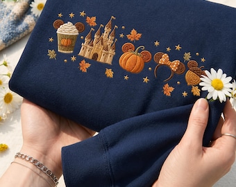 May include: A navy blue sweatshirt featuring embroidered autumn-themed designs including a pumpkin spice drink, a castle, a pumpkin, mouse ears with a bow, and an acorn, all surrounded by small embroidered stars and orange maple leaves.