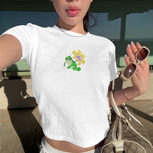 May include: White crop top featuring an embroidered green chameleon holding a yellow and purple flower. The shirt has short sleeves and a crew neck. The design is on the left chest area.