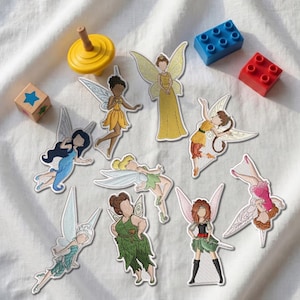May include: A collection of nine embroidered fairy patches featuring diverse figures with wings, alongside a yellow spinning top, a blue star-patterned wooden block, and two plastic building bricks in blue and red colors on a white fabric.