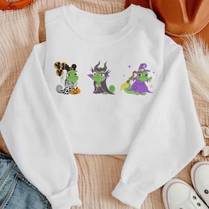 May include: A white long-sleeve sweatshirt featuring three green chameleon illustrations. The first wears a skeleton costume with balloons, the second wears a dark horned robe, and the third wears a purple witch hat and robe holding a broom.