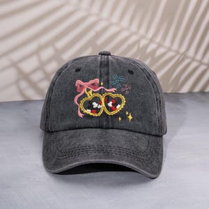 Op de afbeelding: Een donkergrijze baseballcap met een geborduurd ontwerp. Het ontwerp toont twee hartvormige medaillons met Mickey Mouse erin, een roze strik en kleurrijk vuurwerk. De cap heeft een gebogen klep en een stoffen band.