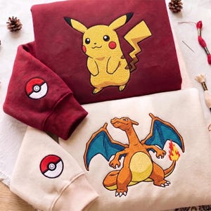 Può includere: Una felpa bordeaux con un Pikachu giallo ricamato e una felpa color crema con un Charizard arancione ricamato. Entrambe le felpe presentano un emblema Poké Ball ricamato sul polsino della manica, con cuciture dettagliate.