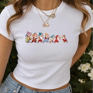 Puede incluir: Una camiseta blanca de manga corta con un estampado gráfico de siete pequeños personajes de dibujos animados en fila, cada uno con ropa y sombreros coloridos. Los personajes aparecen en varias posturas con tonos vibrantes rojos, azules y verdes.