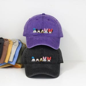 Op de afbeelding: Twee baseball caps, één paars en één zwart, op elkaar gestapeld. Elke cap heeft een geborduurd ontwerp van stripfiguren. De paarse cap heeft een vervaagde uitstraling. Andere caps zijn zichtbaar op de achtergrond.