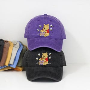 Può includere: Due cappellini da baseball sovrapposti, uno viola e uno nero. Ogni cappellino presenta un'immagine ricamata di Winnie the Pooh che tiene un barattolo di miele, circondato da api. La parola "HUNNY" è sul barattolo di miele.