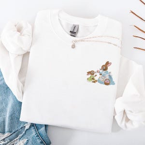 Könnte beinhalten: Weißes Sweatshirt mit Rundhalsausschnitt und gesticktem Design von zwei Hasen. Ein Hase trägt ein blaues Kleid und eine Schürze und hält eine rosa Blume. Der andere Hase trägt ein grünes Kleid. Eine silberne Halskette mit Anhänger ist ebenfalls sichtbar.