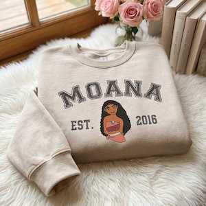 May include: A beige crewneck sweatshirt featuring the text MOANA in a dark grey collegiate font, with the text EST. 2016 on either side of a graphic illustration of the character Moana wearing a pink top and a blue necklace.