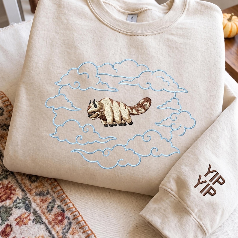 K&ouml;nnte beinhalten: Ein cremefarbenes Sweatshirt mit Rundhalsausschnitt und einer Stickerei eines beigen und braunen fliegenden Bisons, umgeben von hellblau gestickten Wolken. Der rechte &Auml;rmel zeigt den braun gestickten Text YIP YIP in einer serifenlosen Schrift.