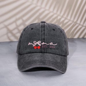 May include: A charcoal gray baseball cap with a curved brim. The cap features the embroidered word "mama" in a cursive font, with a bow and two cherry designs. The text "EST. 2026" is also embroidered on the cap.