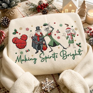 Könnte beinhalten: Ein cremefarbenes Sweatshirt mit einer Stickerei von drei Charakteren aus Nightmare Before Christmas, einem roten Mickey-förmigen Ornament und dem Text "Making Spirits Bright" in grüner Schreibschrift, umgeben von Weihnachtssymbolen.