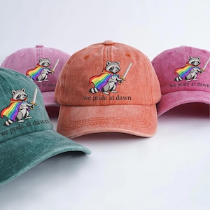 Puede incluir: Varias gorras de béisbol en tonos coral, turquesa y rosa. Cada gorra presenta un mapache bordado con una capa de arcoíris y una espada, con el texto "we pride at dawn".