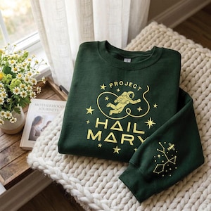 Può includere: Una felpa verde scuro con scritte e grafiche dorate. Il design include un astronauta, stelle e le parole "PROJECT HAIL MARY". La manica presenta un disegno di costellazione. La felpa è piegata su una coperta di maglia color crema.