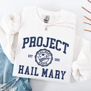 Peut inclure: Sweat-shirt blanc avec texte brodé bleu marine "PROJECT HAIL MARY" et un écusson circulaire. L'écusson comprend les mots "HAIL MARY EARTH" et l'année "2021". Un collier argenté avec un pendentif est posé sur le sweat-shirt.