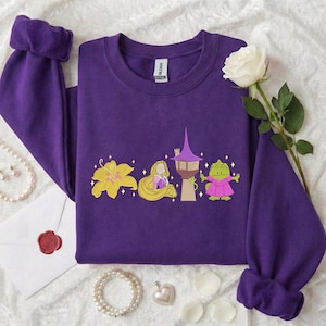 Könnte beinhalten: Ein lila Sweatshirt mit Rundhalsausschnitt und langen Ärmeln. Auf der Vorderseite befinden sich eine gelbe Blume, eine blondhaarige Figur, ein Turm und ein grüner Frosch in einem rosa Kleid. Eine weiße Rose und Schmuck sind im Hintergrund.