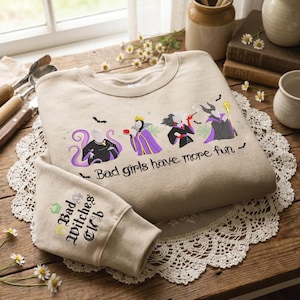 Puede incluir: Una sudadera beige de cuello redondo con un bordado de cuatro villanas y el texto "Bad girls have more fun" y "Bad Witches Club" en la manga. El diseño incluye detalles en morado, negro y verde con pequeños murciélagos.