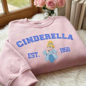 May include: A light pink crewneck sweatshirt featuring embroidered blue text that reads CINDERELLA EST. 1950. Centered below the text is an embroidered graphic of a blonde character wearing a blue dress and matching gloves with a blue headband.