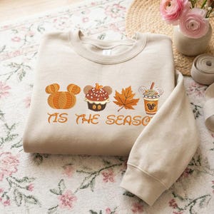 Könnte beinhalten: Ein beigefarbenes Sweatshirt mit Rundhalsausschnitt und einer Stickerei, die einen Kürbis, einen Cupcake, ein Herbstblatt und einen Becher zeigt, alle mit Details im Mickey-Mouse-Stil. Der Text 'TIS THE SEASON' ist in Orange unter den Symbolen gestickt.