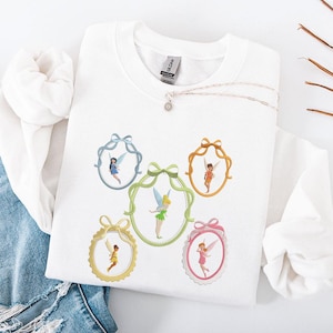 Peut inclure: Sweat-shirt blanc orné de broderies de fées dans des cadres décoratifs. Les cadres sont de différentes couleurs, dont le bleu, le vert, l'or et le rose, chacun avec un nœud. Un collier argenté avec un pendentif est également visible.