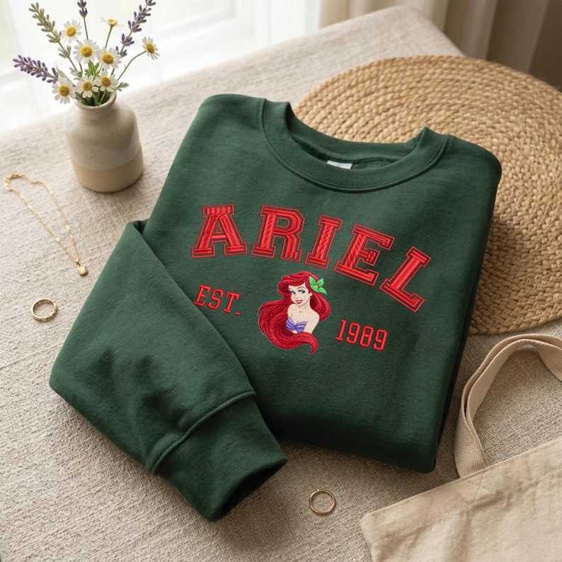 May include: A dark green crewneck sweatshirt featuring red embroidered text that reads ARIEL EST. 1989. Below the text is an embroidered graphic of the character Ariel with long red hair, wearing a purple seashell top and a green hair accessory.