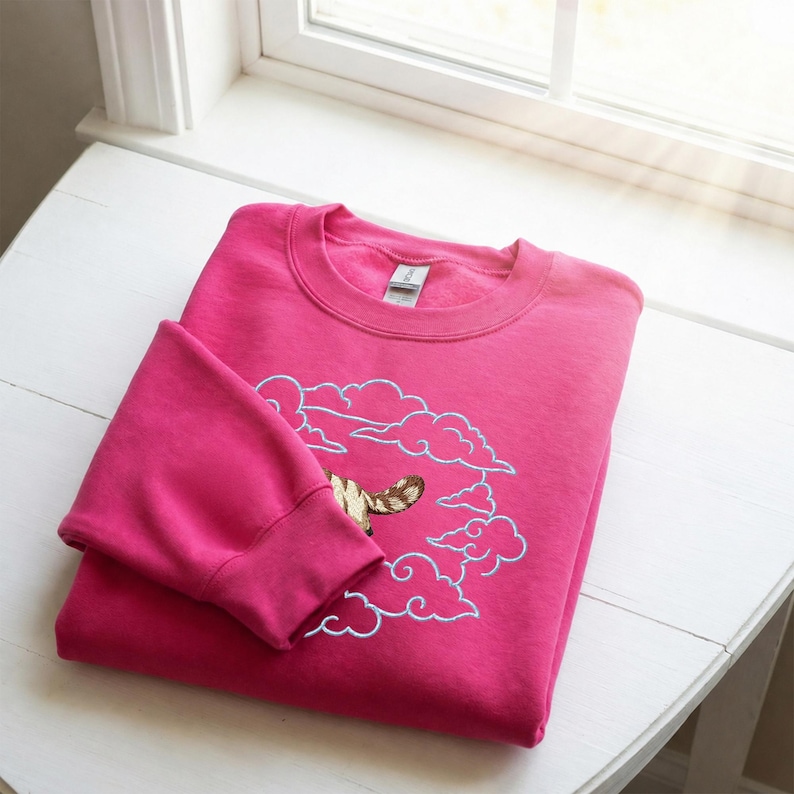 K&ouml;nnte beinhalten: Ein leuchtend pinkfarbenes Sweatshirt mit Rundhalsausschnitt und einer Stickerei, die einen braun-beigen gestreiften Katzenschwanz zeigt, der aus einem Kreis hellblauer gestickter Wolken ragt. Das Kleidungsst&uuml;ck hat lange &Auml;rmel und einen gerippten Kragen.