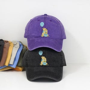 Può includere: Due cappellini da baseball, uno viola e uno nero, impilati l'uno sull'altro. Ogni cappellino presenta un'immagine ricamata di un orso che tiene in mano un palloncino. L'orso è seduto accanto a un vasetto di miele. La parola "Honey" è scritta sul vasetto.