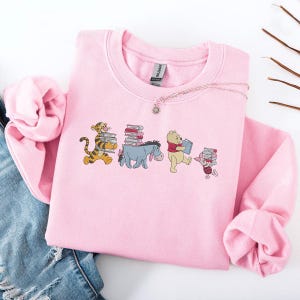 Könnte beinhalten: Hellrosa Sweatshirt mit einem Cartoon-Design von Winnie-the-Pooh-Figuren, die Bücher tragen. Tigger, Eeyore, Winnie Puuh und Ferkel sind abgebildet. Eine Halskette mit einem Sonnenanhänger ist ebenfalls zu sehen.