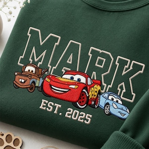 Peut inclure: Un sweat-shirt vert foncé avec le texte brodé MARK EST. 2025. Sous le nom se trouvent trois voitures de dessin animé brodées : une dépanneuse marron, une voiture de course rouge avec le numéro 95 et une voiture de sport bleu clair.