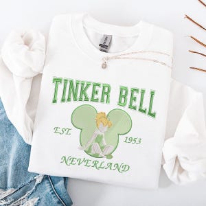 Pode incluir: Um moletom branco com a inscrição "TINKER BELL" em verde. Abaixo, um contorno verde das orelhas do Mickey Mouse com uma imagem bordada de Tinker Bell. O moletom também exibe "EST. 1953 NEVERLAND". Um colar de prata está colocado em cima.