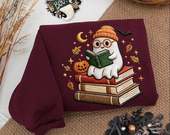 May include: A maroon sweatshirt featuring an embroidered design of a white ghost wearing an orange beanie and round glasses, reading a green book while sitting on a stack of three books, surrounded by a pumpkin, autumn leaves, stars, and a moon.
