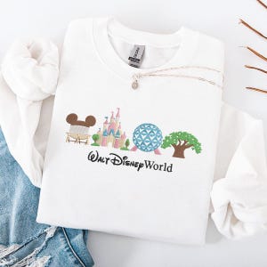 Puede incluir: Una sudadera blanca con un diseño bordado colorido que presenta los emblemáticos monumentos de Walt Disney World. El diseño incluye un helado con forma de Mickey Mouse, un castillo, una esfera geodésica y un árbol. Un collar plateado con un pequeño colgante está drapeado sobre la sudadera.