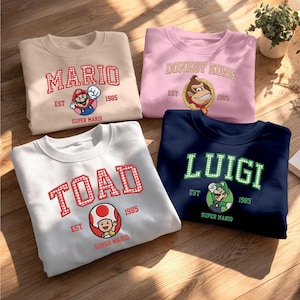 Könnte beinhalten: Vier Rundhals-Sweatshirts mit Nintendo-Charakteren. Ein beiges Shirt mit Mario, ein rosa Shirt mit Donkey Kong, ein weißes Shirt mit Toad und ein dunkelblaues Shirt mit Luigi. Jedes Kleidungsstück zeigt den Namen, EST 1985 und Super Mario Text.