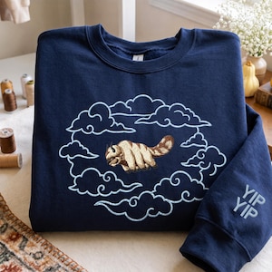 K&ouml;nnte beinhalten: Ein marineblaues Sweatshirt mit Rundhalsausschnitt und einer Stickerei, die ein beiges und braunes pelziges Wesen zeigt, das durch hellblau gestickte Wolken fliegt. Auf dem rechten &Auml;rmel befindet sich der Schriftzug YIP YIP in hellblauer Schrift.