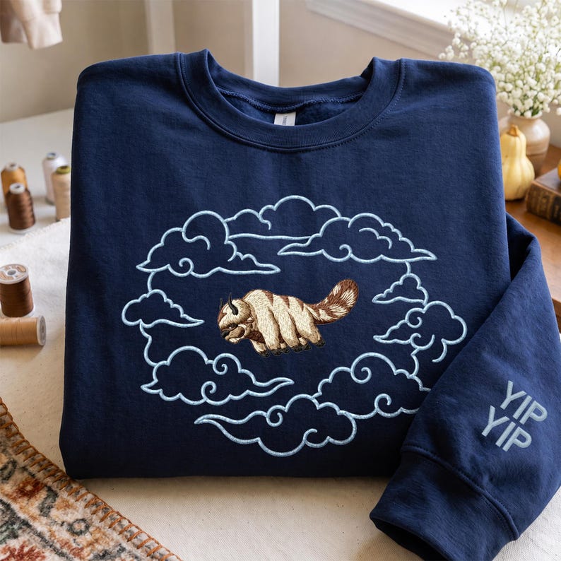K&ouml;nnte beinhalten: Ein marineblaues Sweatshirt mit Rundhalsausschnitt und einer Stickerei, die ein beiges und braunes pelziges Wesen zeigt, das durch hellblau gestickte Wolken fliegt. Auf dem rechten &Auml;rmel befindet sich der Schriftzug YIP YIP in hellblauer Schrift.