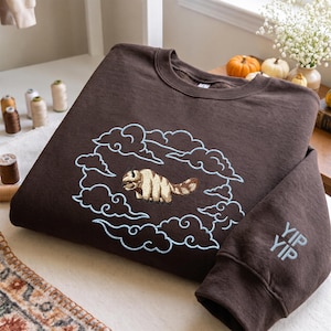 K&ouml;nnte beinhalten: Ein dunkelbraunes Sweatshirt mit Rundhalsausschnitt und einer Stickerei eines beigefarbenen und wei&szlig;en fliegenden Bisons, umgeben von hellblau gestickten Wolken. Der &Auml;rmel ist im unteren Manschettenbereich mit dem Text YIP YIP in hellblauem Garn bestickt.