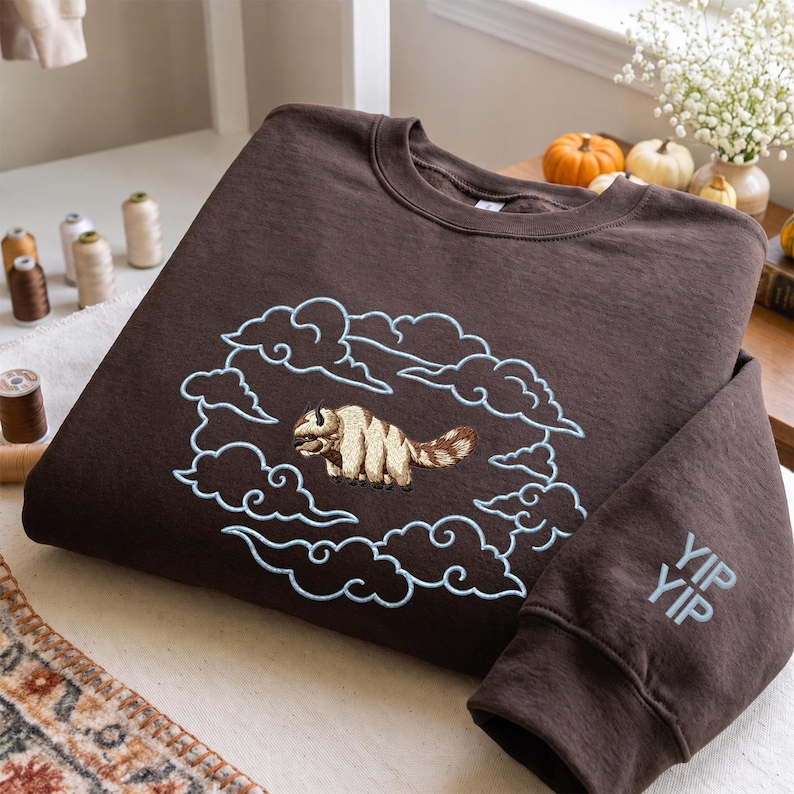K&ouml;nnte beinhalten: Ein dunkelbraunes Sweatshirt mit Rundhalsausschnitt und einer Stickerei eines beigefarbenen und wei&szlig;en fliegenden Bisons, umgeben von hellblau gestickten Wolken. Der &Auml;rmel ist im unteren Manschettenbereich mit dem Text YIP YIP in hellblauem Garn bestickt.