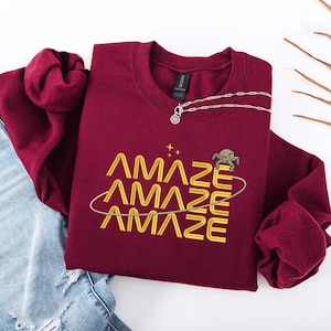 Könnte beinhalten: Ein weinrotes Sweatshirt mit dem Wort "AMAZE" dreimal in Goldfaden gestickt, mit einer Schildkrötengrafik und einer silbernen Halskette. Das Sweatshirt ist auf einer hellen Oberfläche gefaltet.