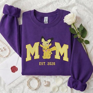 Può includere: Una felpa viola con la scritta "MOM" in lettere gialle, con un design a cartoni animati di Pikachu e Pichu. Il testo "EST. 2026" è in basso. La felpa ha le maniche con i polsini.