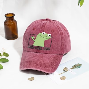 Può includere: Cappellino da baseball bordeaux con finitura invecchiata. Il cappellino presenta una grafica di un lucertola verde dei cartoni animati all'interno di un rettangolo nero, con la scritta "LIZARD LIZARD LIZARD" sotto. Ha una visiera curva e un bottone superiore.