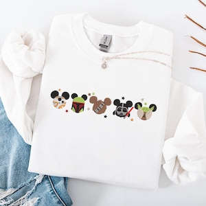 Op de afbeelding: Witte crewneck sweatshirt met geborduurde cartoon karakter ontwerpen. De ontwerpen omvatten karakters met muizenoren, zoals BB-8, Boba Fett, Chewbacca, Darth Vader en Yoda. Een zilveren ketting met een hanger is ook zichtbaar.