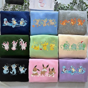 May include: A selection of folded sweatshirts in various colours, each embroidered with different cartoon characters. The sweatshirts are navy blue, light blue, grey, green, light green, beige, black, pink, and purple.