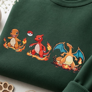 Op de afbeelding: Een donkergroen sweatshirt met een geborduurde afbeelding van drie vuur-wezens in verschillende stadia van evolutie, omringd door vlammen, met een klein rood en wit bal-icoon boven het eerste wezen aan de linkerkant.