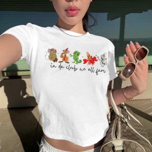 Op de afbeelding: Een wit T-shirt met korte mouwen en een grafische print van vijf geanimeerde figuren op een rij, waaronder een muis, een hagedis, een krab en een sneeuwpop. Onder de figuren staat de tekst "in da clerb we all fam" in zwarte letters.