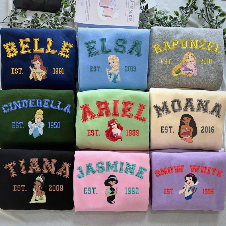 May include: A collection of nine crewneck sweatshirts in various colors, each featuring an embroidered name of a Disney character, a corresponding character illustration, and an establishment year, including Belle, Elsa, Rapunzel, Cinderella, Ariel, Moana, Tiana, Jasmine, and Snow White.