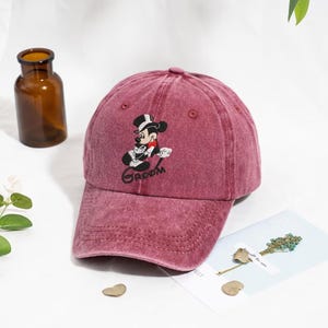 Puede incluir: Gorra de béisbol granate con un diseño bordado de Mickey Mouse con esmoquin y la palabra "Groom" debajo. La gorra tiene una visera curva y un aspecto desgastado, lo que le da un estilo vintage.