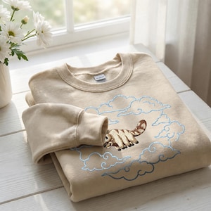 K&ouml;nnte beinhalten: Ein beigefarbenes Sweatshirt mit Rundhalsausschnitt und einer Stickerei, die ein sechsbeiniges, braun gestreiftes Wesen zeigt, das durch einen Kreis aus hellblau gestickten Wolken fliegt. Das Kleidungsst&uuml;ck hat einen gerippten Kragen und lange &Auml;rmel.
