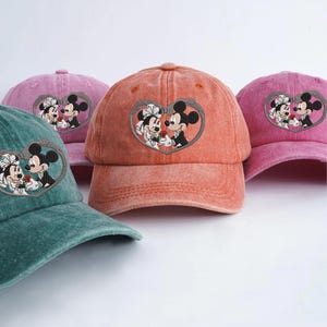 Könnte beinhalten: Eine Auswahl an Baseballkappen in verschiedenen Farben, darunter Türkis, Rosa und Orange. Jede Kappe zeigt ein gesticktes herzförmiges Design mit Mickey und Minnie Mouse in Hochzeitskleidung. Die Kappen haben einen Vintage-Look.