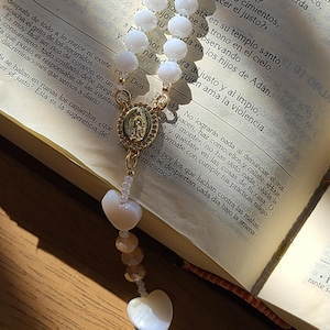 Könnte beinhalten: Ein Rosenkranz mit weißen, facettierten Perlen und herzförmigen Anhängern. Das goldfarbene Mittelstück zeigt ein religiöses Emblem. Der Rosenkranz liegt auf einem offenen Buch mit Text.